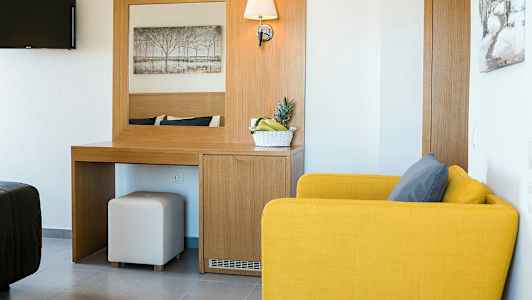 Et hotellrom med en gul lenestol, et skrivebord med speil og en skål med frukt. (AI-generert tekst)