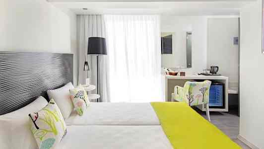 Et lyst hotellrom med to enkeltsenger, dekorative puter og et limegrønt sengeteppe, samt en sitteplass og et skrivebord med speil. (AI-generert tekst)