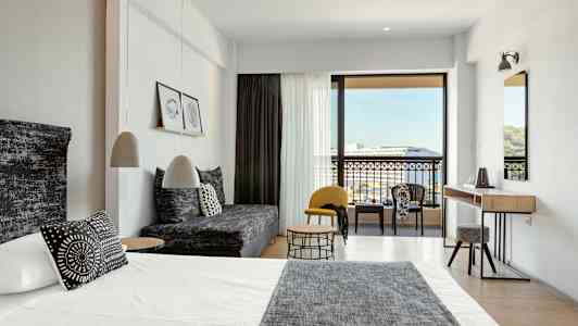 Moderne hotellrom med seng, sofa og sittegruppe samt utsikt mot balkong og omgivelser. (AI-generert tekst)