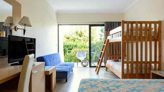 Et hotellrom med køyeseng, blå sofa, skrivebord, TV og balkong med utsikt mot grøntområder. (AI-generert tekst)