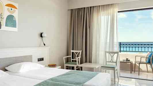 Hotellrom med dobbeltseng, to stoler og et bord ved vindu med havutsikt. (AI-generert tekst)