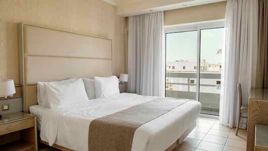 Et hotellrom med en dobbeltseng kledd i hvitt sengetøy, nattbord og store vinduer med gardiner som slipper inn lys. (AI-generert tekst)