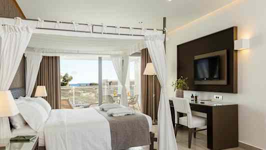 Et elegant hotellrom med en himmelseng, skrivebord og utsikt mot havet fra en balkong. (AI-generert tekst)