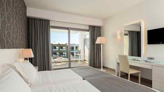 Et hotellrom med to enkeltsenger, skrivebord, stol og en vindusutsikt mot en balkong med bord og stoler. (AI-generert tekst)