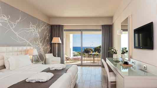 Et hotellrom med to enkeltsenger, skrivebord, sitteplass og vindu mot en balkong med havutsikt. (AI-generert tekst)