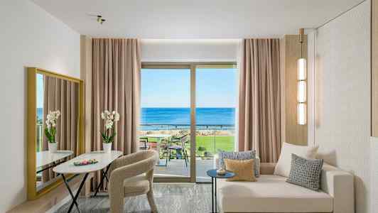 En moderne hotellsuite med sofa, bord, stoler og utsikt over havet gjennom et stort vindu. (AI-generert tekst)