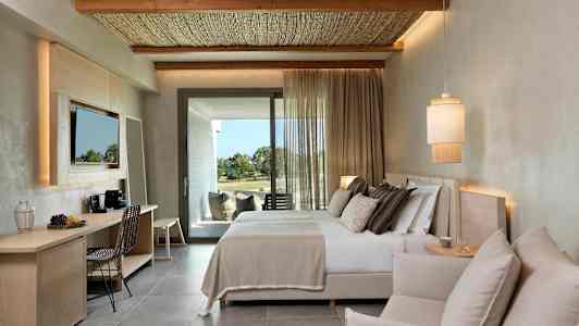 Et moderne hotellrom med en dobbeltseng, en sittegruppe og et skrivebord. Tyg slipper inn dagslys fra balkongdøren. (AI-generert tekst)