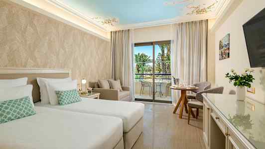 Et hotellrom med to senger, sittegruppe og balkong med utsikt mot palmer. (AI-generert tekst)