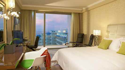 Hotellrom med en dobbeltseng, stoler og utsikt over balkonger og havet gjennom store vinduer. (AI-generert tekst)