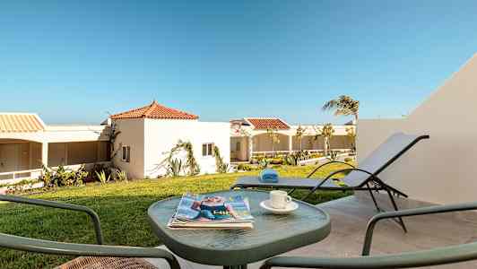 En terrasse med utemøbler, blader og en kopp kaffe, med utsikt mot hvitkalkede hus med røde tegltak og grønne omgivelser under en klar blå himmel. (AI-generert tekst)