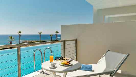 Balkong med utsikt over et basseng og havet i bakgrunnen, et bord med frokost og en solseng. (AI-generert tekst)