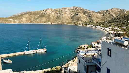 Utsikt over en kyst med små bygninger og fjell i bakgrunnen. Båter ligger fortøyd ved brygga i det klare vannet. (AI-generert tekst)