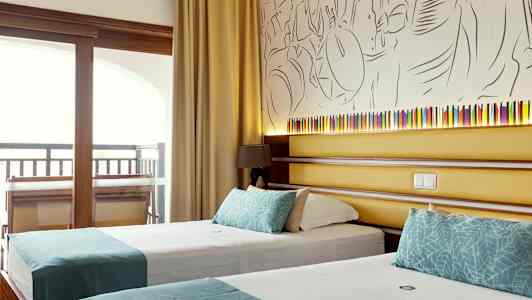 Et hotellrom som viser to enkeltsenger med turkise dekorasjoner, bak sengene er det en vegg med kunstnerisk dekorasjon og en liten lampe. (AI-generert tekst)