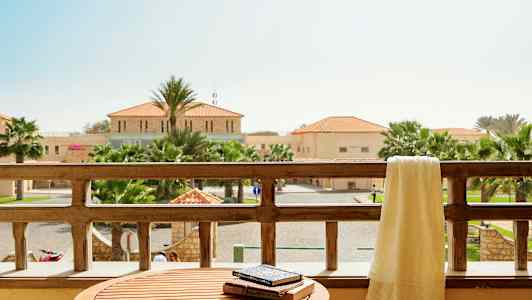 Utsikt fra en balkong med et bord og et håndkle over balkongrekkverket, omkringliggende bygninger og palmer er synlige i bakgrunnen. (AI-generert tekst)