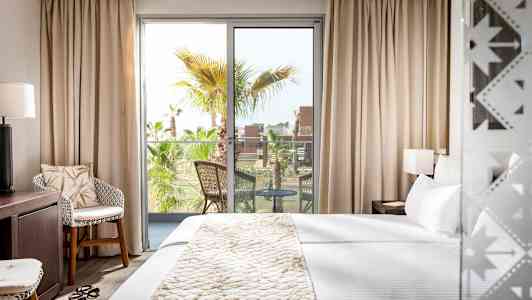 Et hotellrom med en stor seng, en sittegruppe og en balkong med utsikt over palmer. (AI-generert tekst)