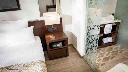 Et hotellrom med en seng, et nattbord med lamper og et bad med glassdør. (AI-generert tekst)