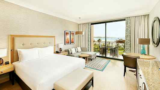 Et hotellrom med en stor seng, sittegruppe, skrivebord og en uteplass som vender mot sjøen. (AI-generert tekst)