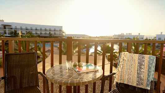 En balkong med to stoler, et bord og utsikt over et basseng og hotellbygninger. (AI-generert tekst)