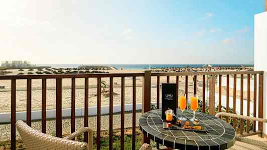 En balkong med utsikt over en strand og havet, med to stoler og et bord dekorert med drikke og snacks. (AI-generert tekst)