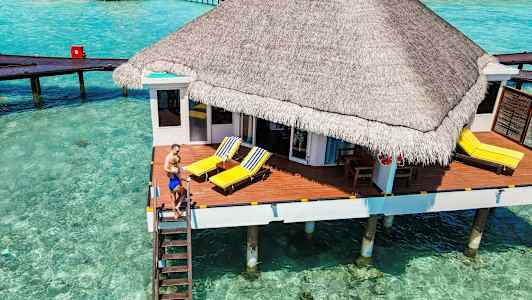 Bungalow på påler over klart turkis vann med solterrasse og strandstoler. (AI-generert tekst)