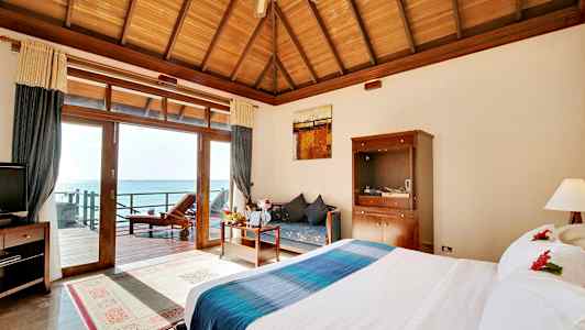 Et hotellrom med en stor seng, et trebord og en sofa, utsikt over havet gjennom glassdører. (AI-generert tekst)