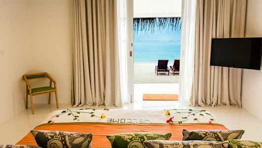 Utsikt fra hotellrom med dekorert seng, ryggstølstol og TV, mot strand og hav gjennom glassdør. (AI-generert tekst)