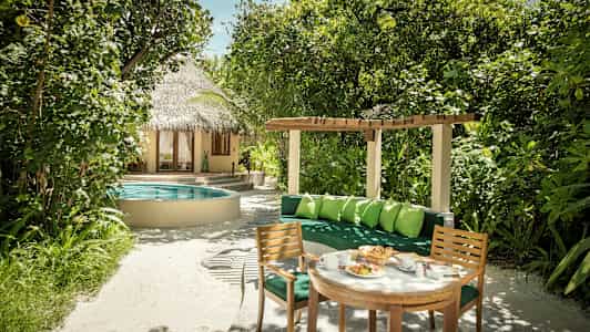 En hage med frodige planter, et rundt bassengområde, et hus med stråtak, og en sittegruppe med bord og stoler. (AI-generert tekst)