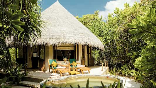 Bilde av en bungalow med stråtak, omgitt av frodig vegetasjon og med en liten basseng i forgrunnen. (AI-generert tekst)