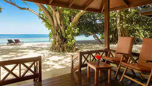 Utsikt fra en veranda med stoler og bord mot en strand med solsenger og hav. (AI-generert tekst)