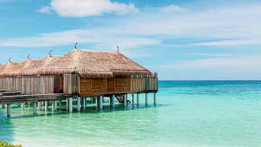 Vannbungalower med stråtak på pæler over turkist vann og klar himmel. (AI-generert tekst)