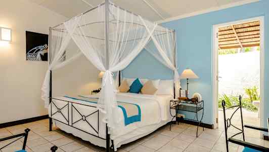 Et soverom med en himmelseng med hvite og blå tekstiler, to nattbord med lamper og en dør som fører til et utendørsområde. (AI-generert tekst)