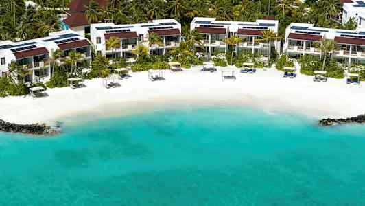 En strand med turkist vann, hvit sand og solsenger foran et boligområde omgitt av palmer. (AI-generert tekst)