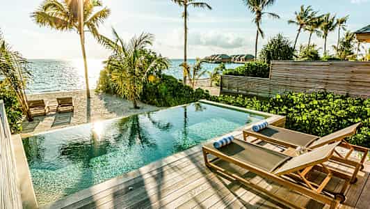 Et basseng med solsenger på en terrasse ved en sandstrand og havet. (AI-generert tekst)