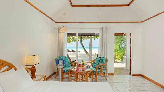 Et hotellrom med seng, to stoler og bord viser utsikt mot en strand gjennom glassdører. (AI-generert tekst)