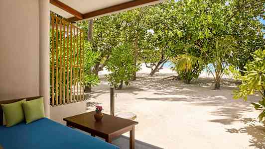 Utsikt fra en terrasse med en blå sofa og et bord med en vase, omgitt av sand og frodige trær nær sjøen. (AI-generert tekst)