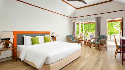 Et rom med en komfortabel dobbeltseng, sittegruppe med stoler og et bord, og store vinduer som gir utsikt mot en frodig hage. (AI-generert tekst)