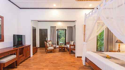 Et rom med tremøbler, en seng med myggnetting og en sittegruppe med utsikt mot en hage. (AI-generert tekst)