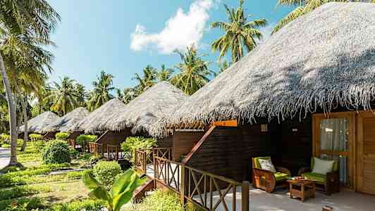 Bildet viser flere bungalower med stråtak omgitt av tropiske planter og palmer. (AI-generert tekst)