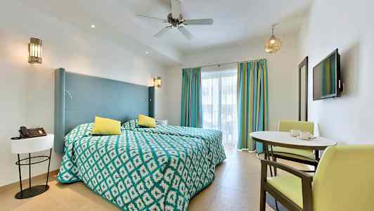 Et hotellrom med to enkeltsenger, en liten spiseplass og en vegg-TV foran vinduer med fargerike gardiner. (AI-generert tekst)