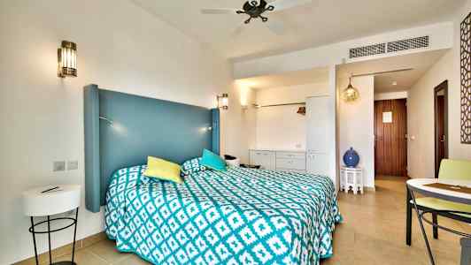 Et hotellrom med en dobbeltseng, blått sengegavl, fargerike puter og et mønstret teppe. Detaljer inkluderer møbler og dekorative elementer. (AI-generert tekst)