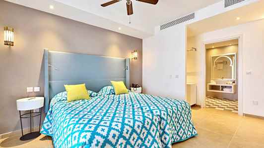 Et hotellrom med en seng som har et blått og hvitt mønstret sengeteppe og gule puter. I bakgrunnen er et baderom. (AI-generert tekst)