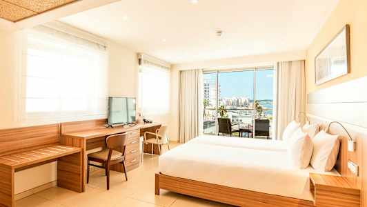 Et lyst hotellrom med en stor seng, skrivebord og TV, samt en balkong med utsikt over en by og havet. (AI-generert tekst)