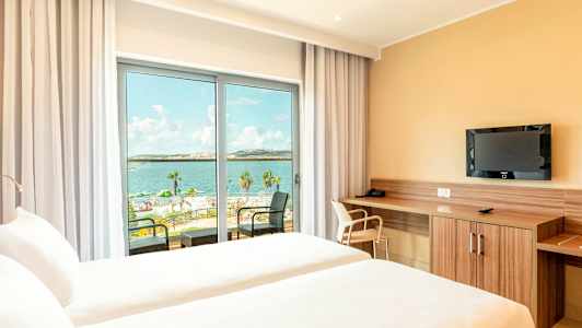 Et hotellrom med to senger og utsikt over havet gjennom en glassdør. (AI-generert tekst)