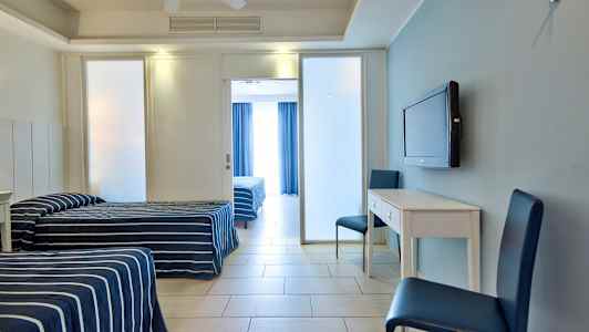 To enkeltsenger med stripete blå sengetepper, et hvitt skrivebord med to blå stoler og en veggmontert TV, samt en glassdør til rommet bak. (AI-generert tekst)