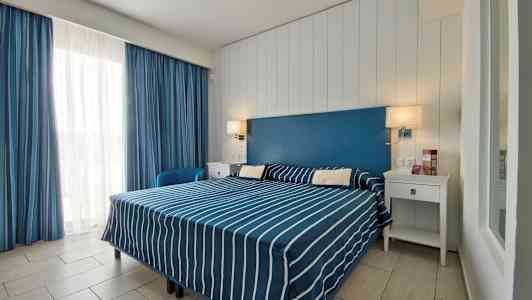 Et hotellrom med en dobbeltseng med blå- og hvitstripete sengetøy, en blå sengegavl og hvite møbler. (AI-generert tekst)