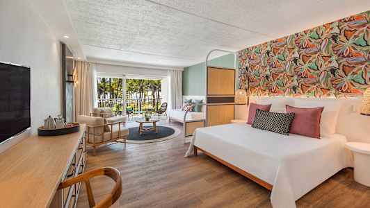 Hotellrom med dobbeltseng, sittegruppe, skrivebord og balkong med utsikt over palmer og hage. (AI-generert tekst)