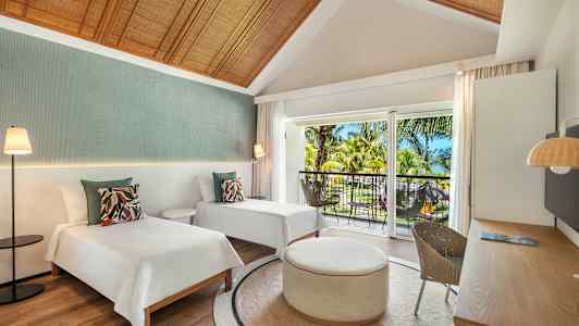 Et hotellrom med to enkeltsenger, et rundt bord og en balkong med utsikt over tropiske trær. (AI-generert tekst)