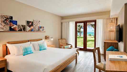 Et hotellrom med en dobbeltseng, tremøbler, og en glassdør som åpner opp til en grønn hage og havutsikt. (AI-generert tekst)