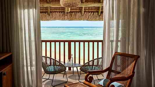 Utsikt over havet fra et rom med en overbygd balkong. Mellom gjennomsiktige gardiner er det to hagestoler ved et bord. (AI-generert tekst)