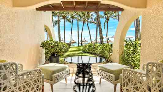 Utsikt fra en uteplass med stoler og et bord mot en hage med palmer og strand i bakgrunnen. (AI-generert tekst)